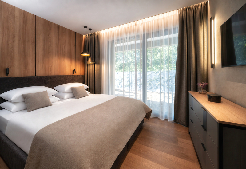 Modern hotel room with king bed, wood-paneled headboard, layered pillows, pendant bedside lamps, sheer floor-to-ceiling curtains and dresser