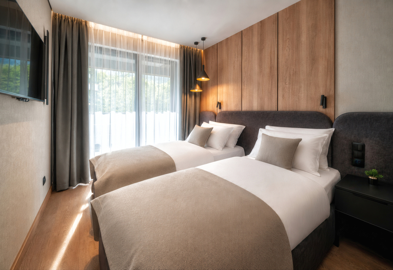 Twin beds with plush headboards, wood-paneled wall, pendant bedside lamps, sheer-curtained window and TV
