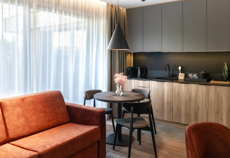 Modern suite kitchenette with wood cabinets, black sink and coffee machine, round dining table, vase of flowers and leather sofa.