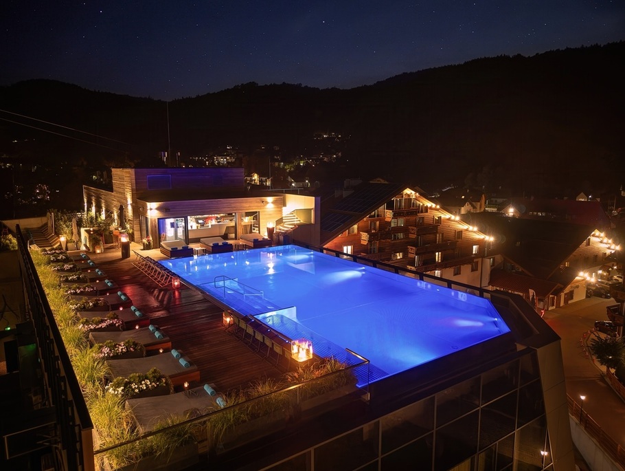Rooftop-Infinity-Pool nachts blau beleuchtet, Holzdeck, Liegen, Barbereich und Blick auf ein Bergdorf.