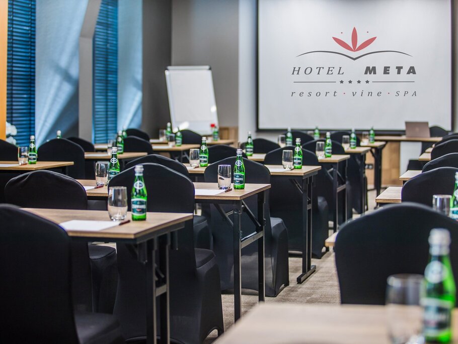 Konferenzraum in Schulungsanordnung, Wasserflaschen und Gläser auf Tischen, Leinwand mit Hotel META-Logo und Flipchart