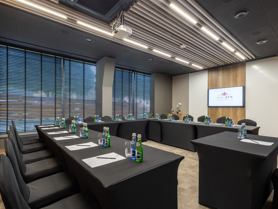 Konferenzraum in U‑Form mit schwarzen Tischdecken, Mineralwasser und Schreibsets, Deckenprojektor, Wandmonitor mit Hotel-Logo und modernen Jalousien.