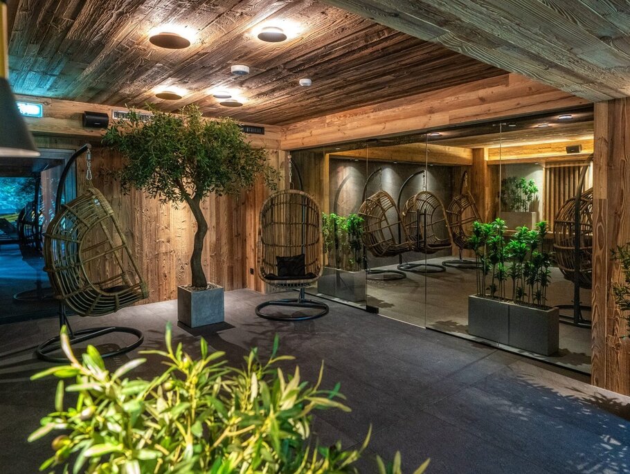 Holzverkleidete Relax-Lounge mit hängenden Rattan-Schaukelstühlen, Topfbäumen, warmem Deckenlicht und Glaswand, die die gemütliche Spa-Sitzgruppe spiegelt.
