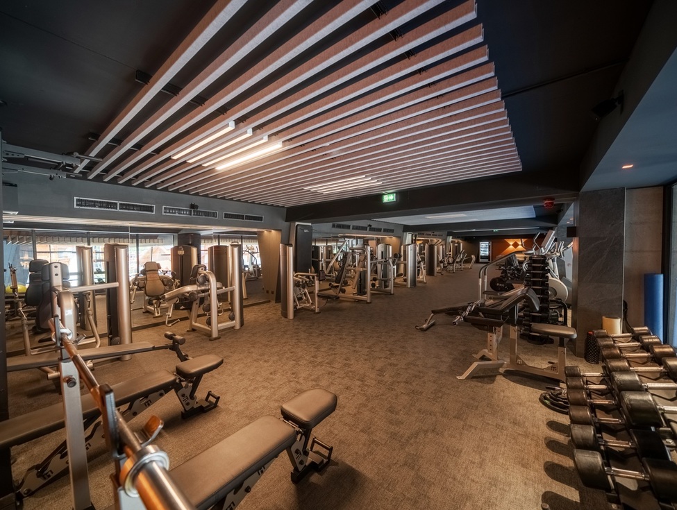 Geräumiger Hotel-Fitnessraum mit Kraftgeräten, Hanteln, Bänken, Spiegelwand und warmem Licht unter Holzlamellen-Decke