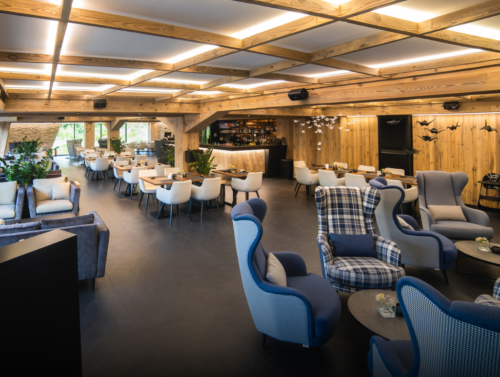 Cozy hotel lounge with wooden-beam ceiling, bar, white dining chairs, blue plaid armchairs, plants and warm lighting.