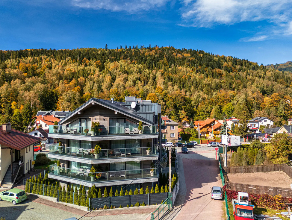 Modernes Chalet-Gästehaus mit Glasbalkonen und möblierten Terrassen, Blick auf herbstlich bewaldete Hügel und Dorf.