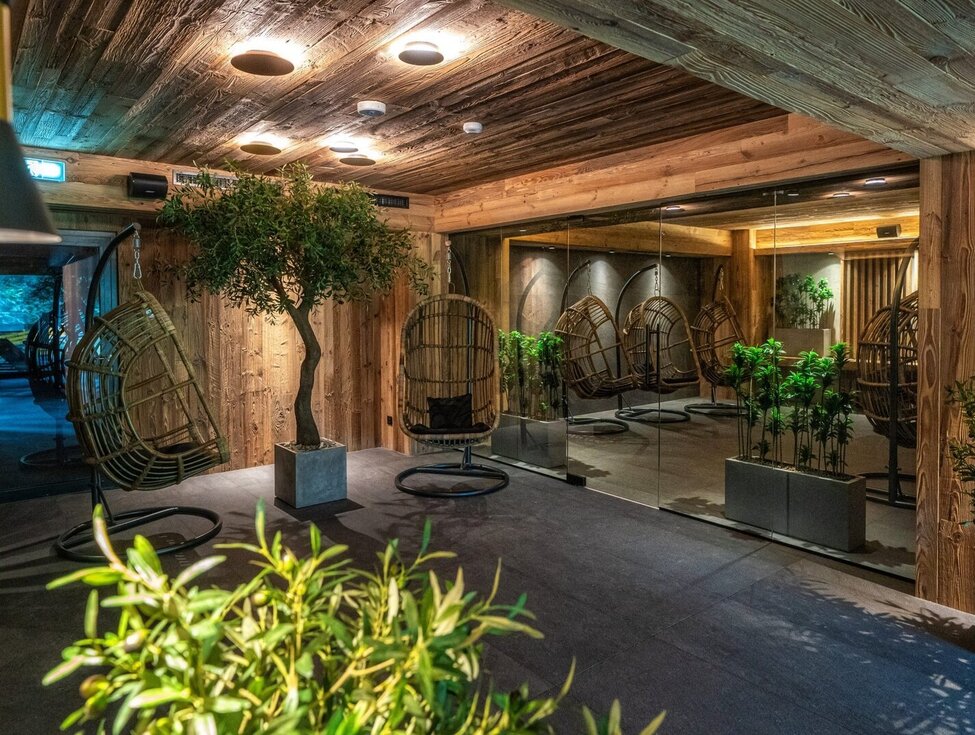 Gemütlicher Spa-Relaxbereich mit hängenden Rattan-Schaukelstühlen, warmen Holzverkleidungen, grünen Pflanzen und Glaswand.