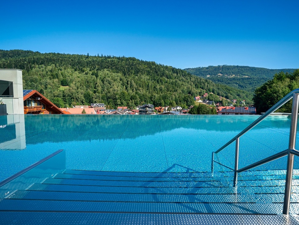 Rooftop infinity pool with steps and stainless handrail, overlooking a forested mountain valley and village under clear blue sky