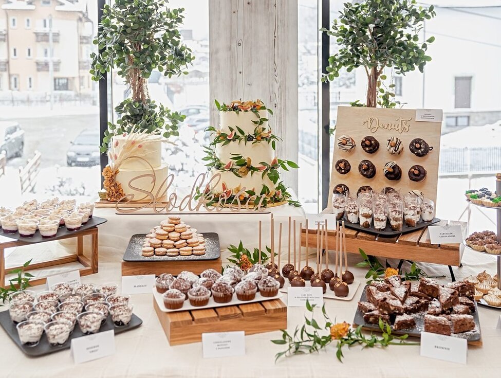 Elegantes Dessertbuffet mit mehrstöckiger Hochzeitstorte, Donut-Wand, Muffins, Brownies und Cakepops am Fenster mit verschneiter Aussicht