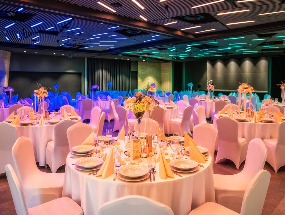 Hotel banquet hall with round tables, white chair covers, floral centerpieces and folded napkins beneath blue-green LED ceiling lights.