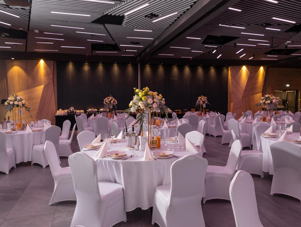 Geräumiger moderner Bankettsaal für Empfang: runde Tische mit weißen Tischdecken, hohe Blumenarrangements, gefaltete Servietten, Buffet und stimmungsvolle Beleuchtung