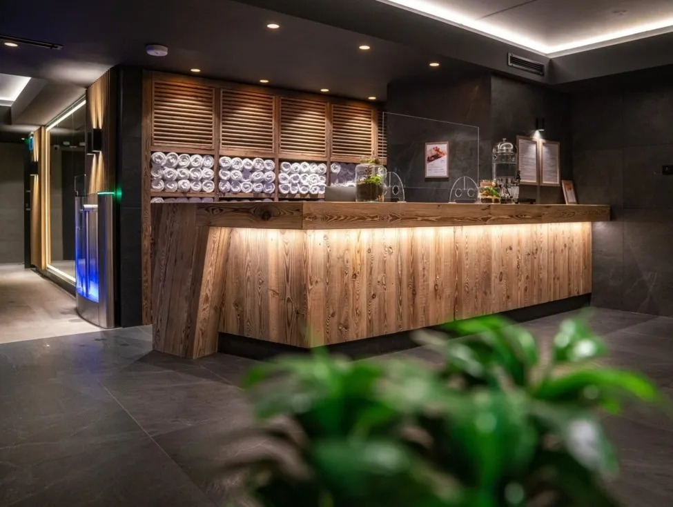 Hotel spa reception with wooden front desk, backlit counters, shelves of rolled white towels and soft ambient lighting