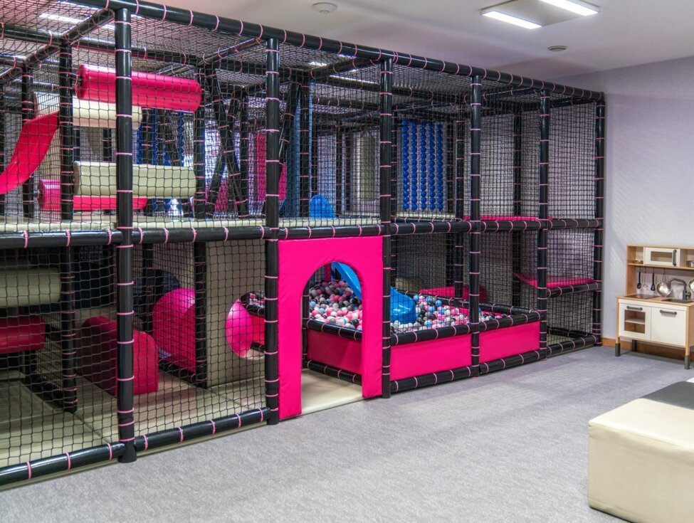 Hotel indoor kids' playroom with multi-level soft-play structure, pink ball pit, slide and tunnels, plus a children's play kitchen