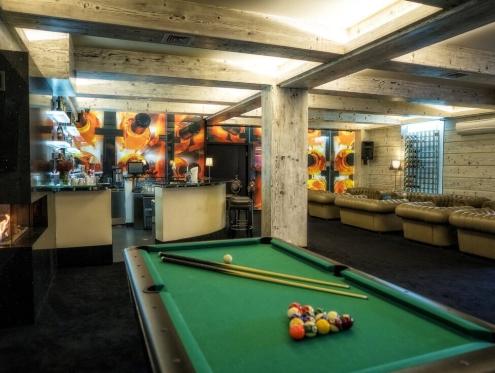 Stylish hotel game lounge with green pool table, bar counter, tufted leather sofas, fireplace, wood‑beamed ceiling and vibrant mural.