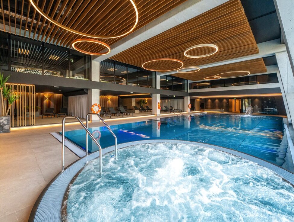 Innen-Spa mit sprudelndem Whirlpool im Vordergrund, großem Schwimmbad, Holzlamellen-Decke und runden LED-Ringen