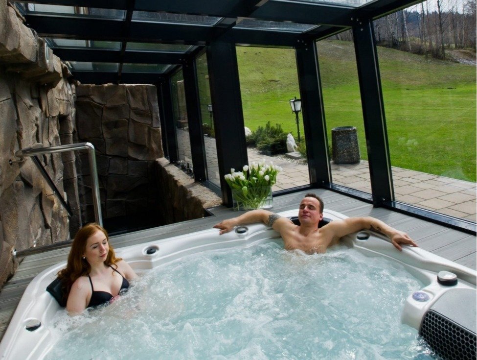 Paar entspannt im privaten, verglasten Whirlpool mit Sprudelfunktion, Tulpen auf der Terrasse und Blick auf grünen Rasen.