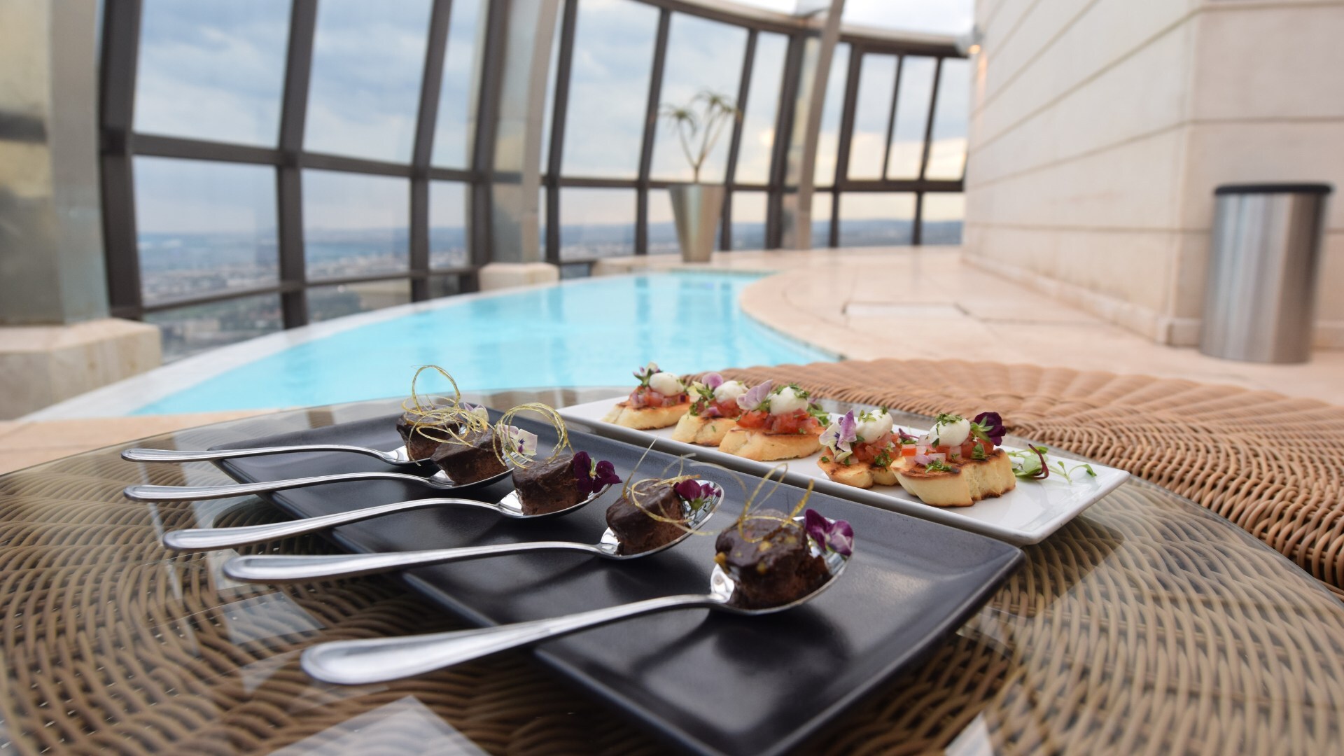 Dachpool mit bodentiefen Fenstern und Stadtblick; Canapés und Schokoladenhäppchen auf Glas­tisch neben dem Pool.