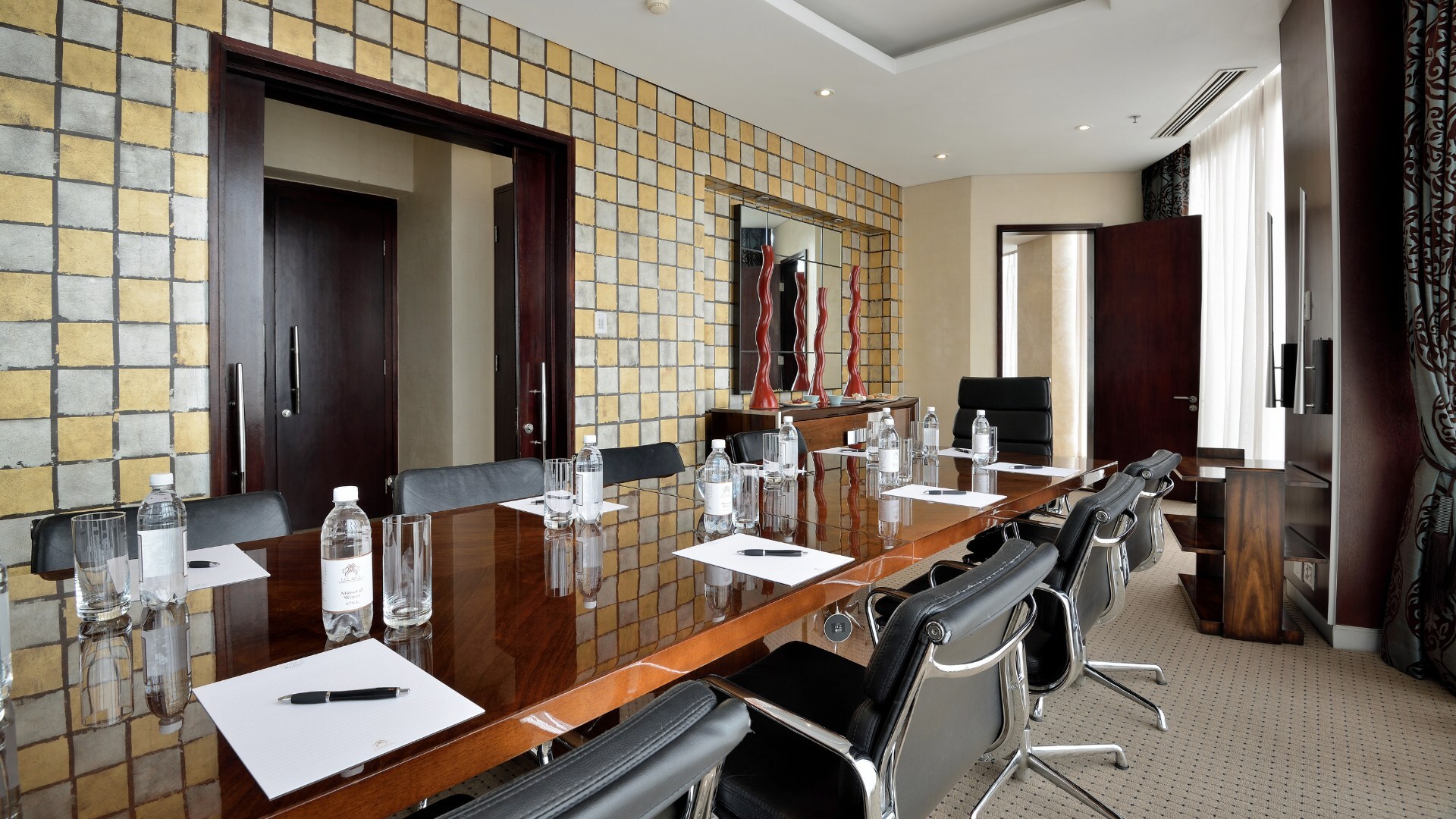 Konferenzraum mit glänzendem Holztisch, Lederdrehstühlen, Wasserflaschen, Schreibblöcken, Kachelwand und Tageslicht.