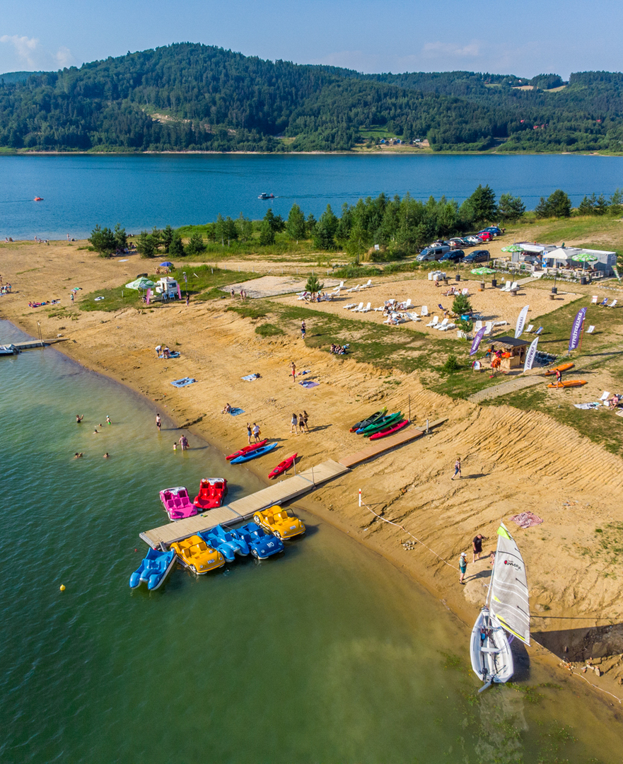 Plaża z wypożyczalnią kajaków, rowerków wodnych i żaglówką nad jeziorem z lasami w tle, goście korzystają z kąpieli i plażowania