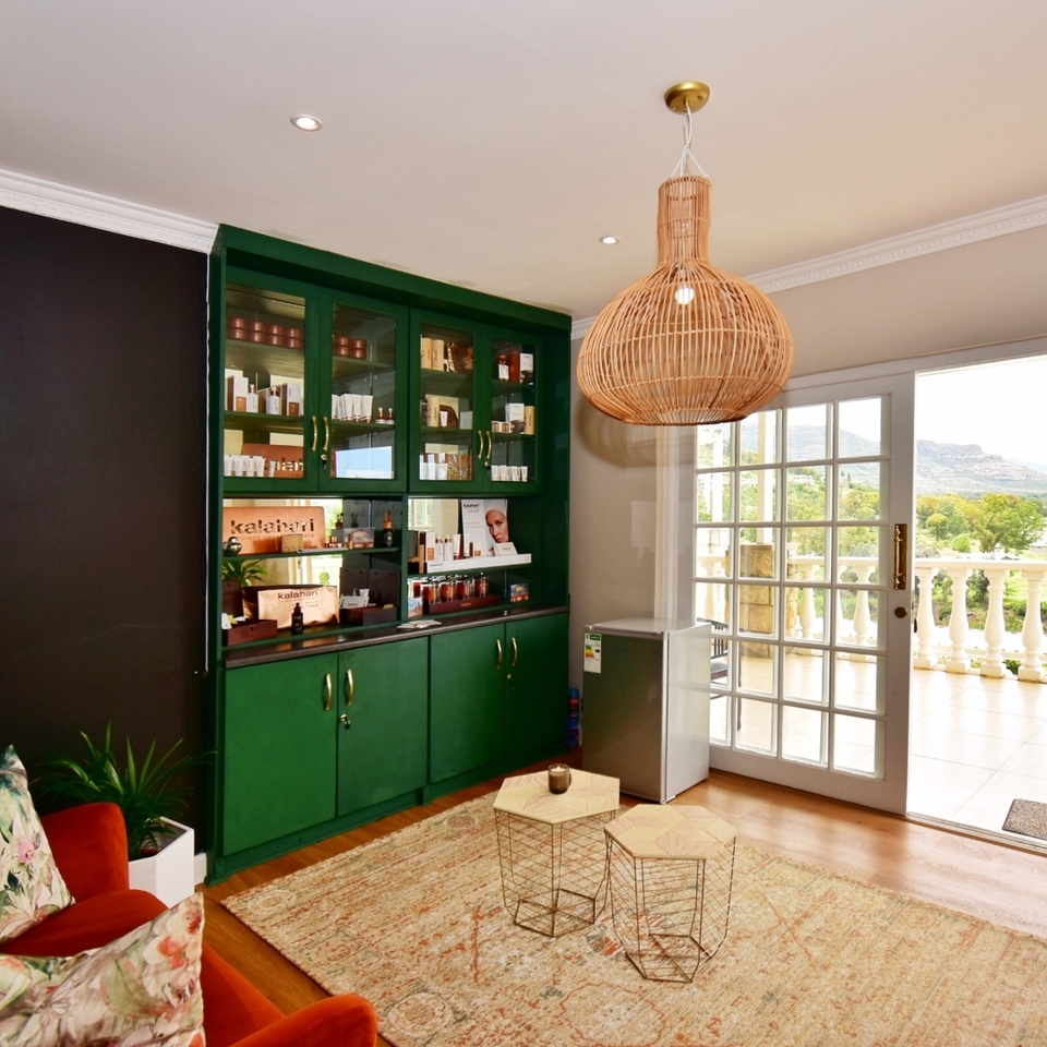 Cozy spa lounge with green display cabinets of products, rattan pendant, velvet sofa, and balcony overlooking mountains.