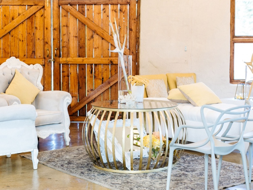 Cozy lounge with tufted white armchairs, yellow cushions, gold round coffee table, bar cart, rustic barn doors, bright window