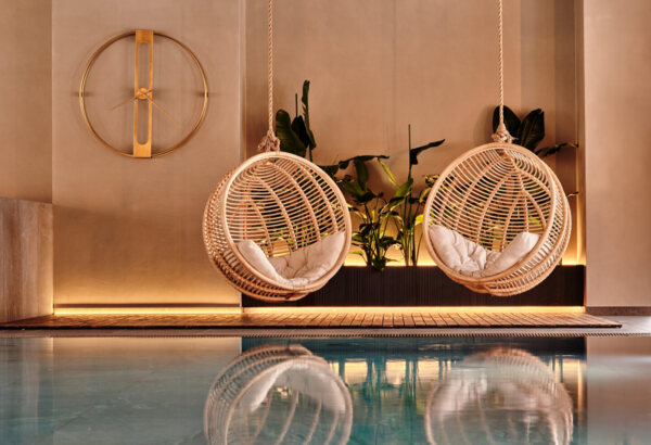 Moderní závěsné ratanové křesla s polštáři u vnitřního bazénu, doplněné zelenými rostlinami a stylovými hodinami na zdi.