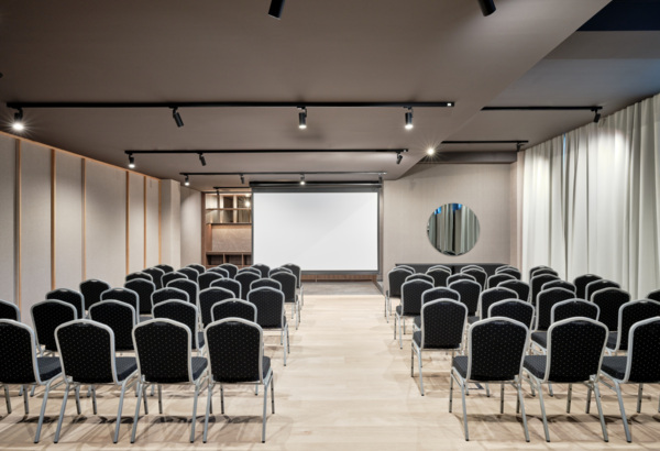 Nowoczesna sala konferencyjna z rzędem czarnych krzeseł, jasną podłogą i ekranem projekcyjnym do prezentacji