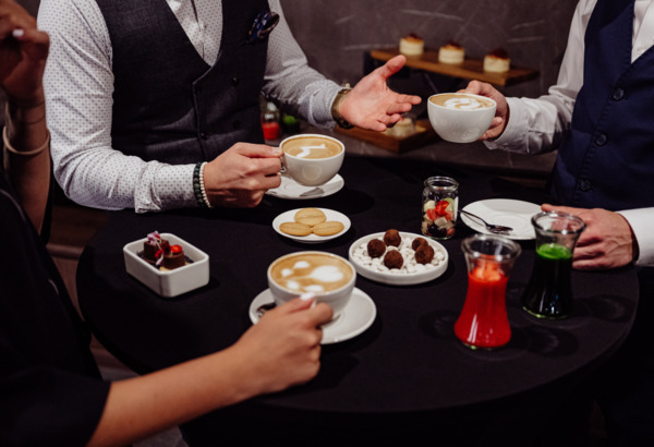 Goście w eleganckich strojach piją kawę latte przy stole z deserami i sokami w stylowej kawiarni hotelowej.