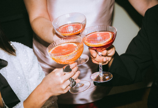 Trzy osoby w eleganckich strojach stukają się kieliszkami koktajli z plasterkiem pomarańczy
