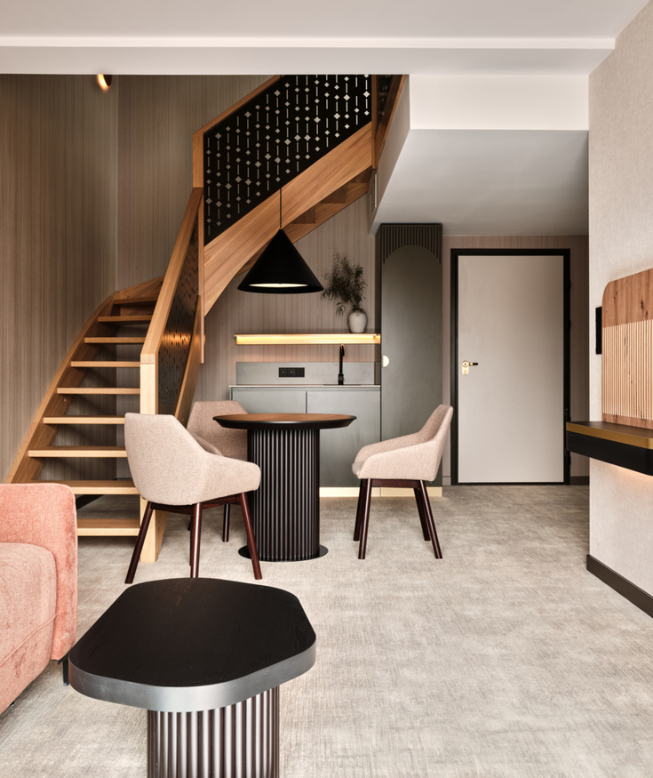 Nowoczesny pokój hotelowy z drewnianymi schodami, miękkim różowym kanapą, jadalnią z dwoma krzesłami i dużym telewizorem na ścianie.