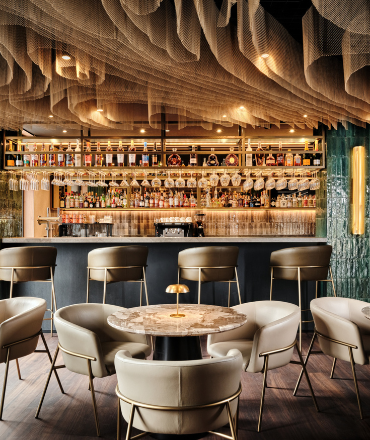 Stylový hotelový bar s mramorovými stoly a pohodlnými béžovými křesly, zelené dlaždice a umělecký strop.