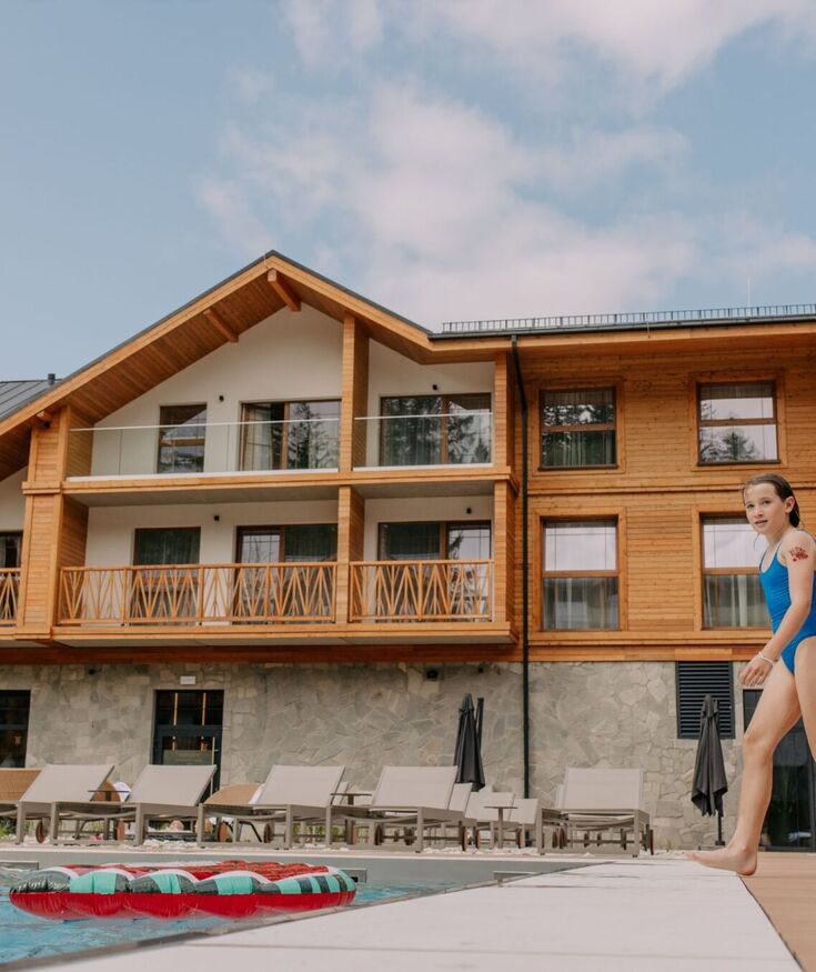 Drewniany hotel z balkonami i basenem, z leżakami przy basenie, dziecko w stroju kąpielowym spaceruje po tarasie