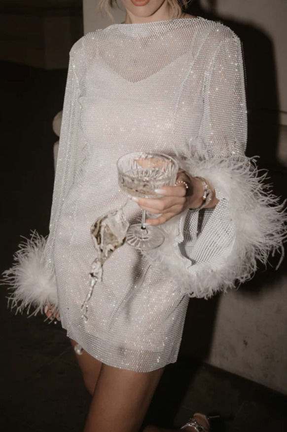 Kobieta w połyskującej sukience z piórami na rękawach, trzyma kieliszek z rozlewającym się drinkiem na tle przytulnego wnętrza