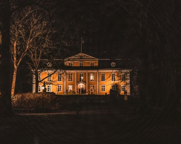 Valoilla korostettu historiallinen kartano, jossa klassista arkkitehtuuria ja metsää ympärillä yöllä, kutsuva sisäänkäynti