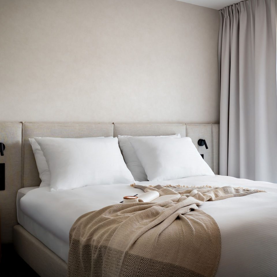 Elegancki hotelowy pokój z podwójnym łóżkiem, białą pościelą, beżowym pledem, tapicerowanym wezgłowiem, drewnianym stolikiem i lampkami
