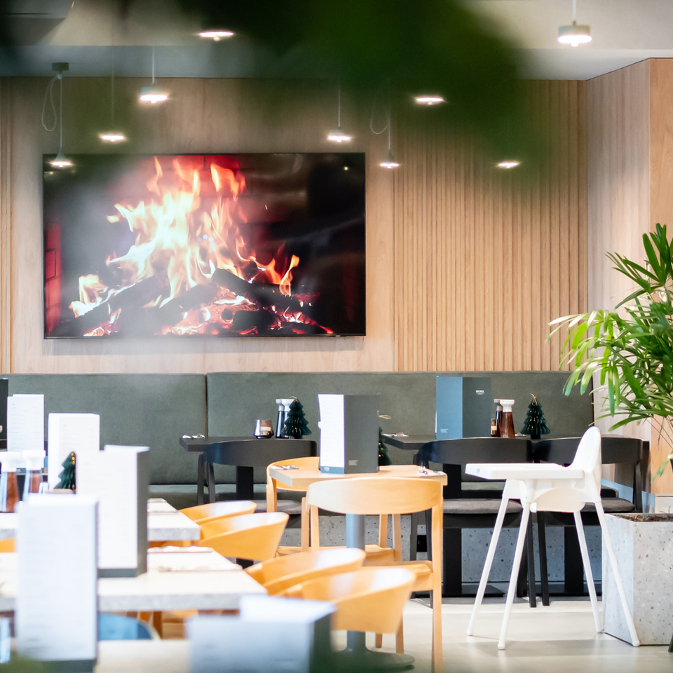 Modern hotel dining area with wood-paneled wall, large screen showing a fireplace, pendant lights, mixed seating and abundant plants