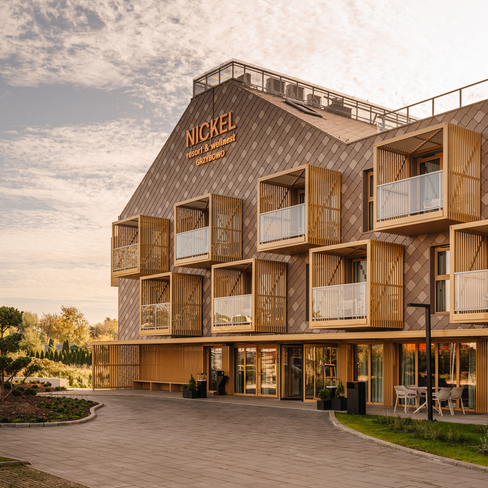 NICKEL resort & wellnest — nowoczesna fasada hotelu wellness z drewnianymi balkonami, szklanymi balustradami, zielonym wejściem i zewnętrznym miejscem do siedzenia.