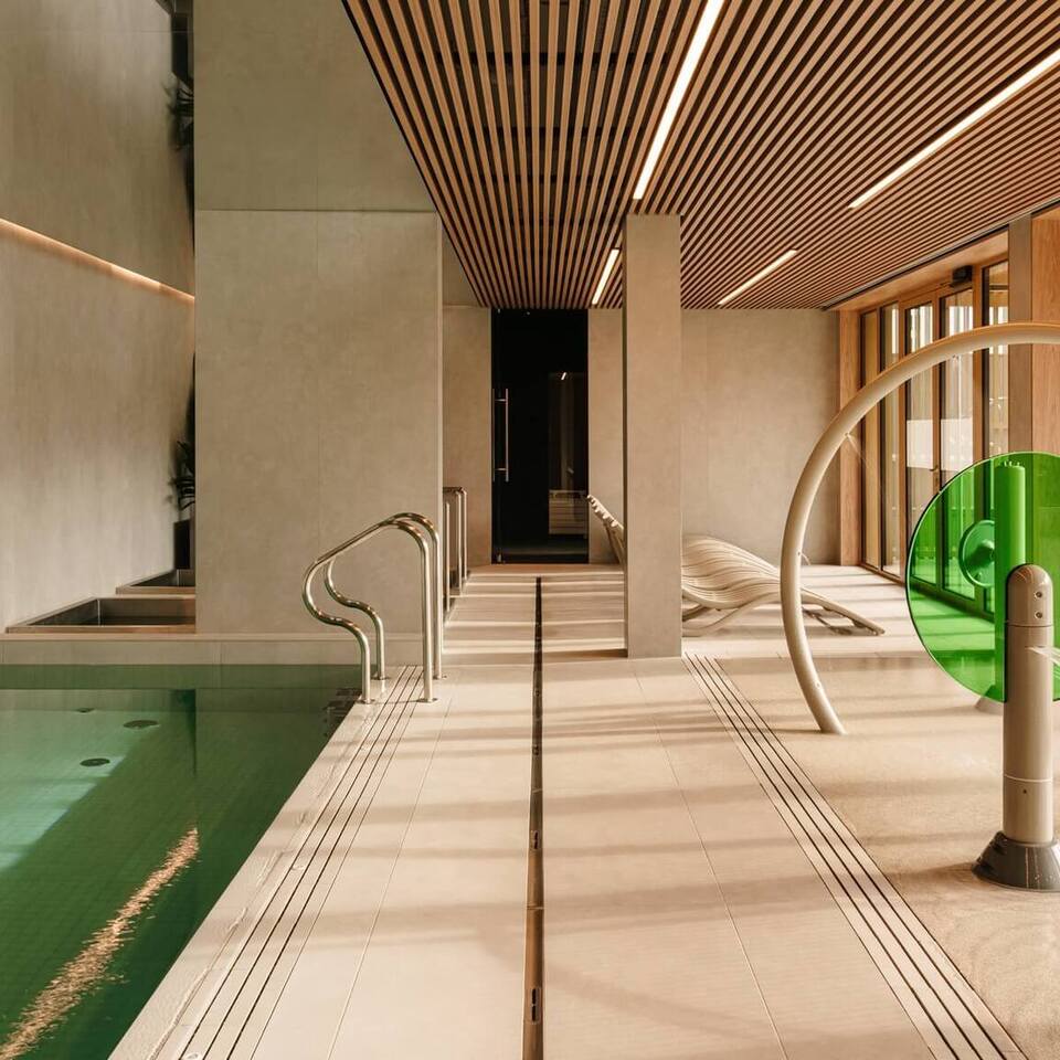 Wewnętrzne spa z zielonym basenem w płytkach, drewnianym listwowanym sufitem, oknami od podłogi do sufitu, leżakami i okrągłą fontanną.