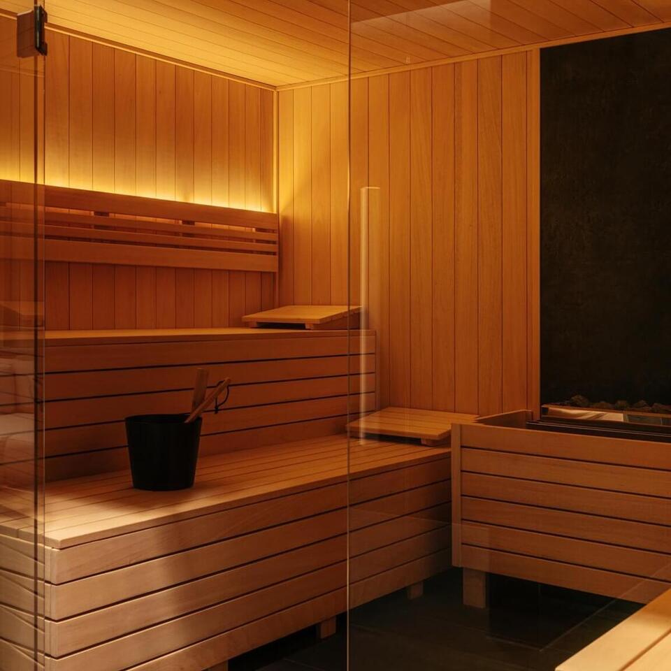 Hotelowa sauna obłożona drewnem, wielopoziomowe ławki, szklane drzwi, piec z kamieniami, wiadro i ciepłe oświetlenie