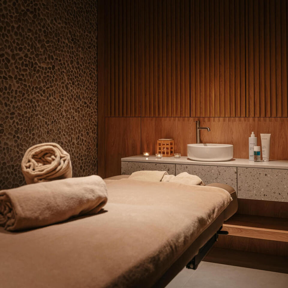 Spokojny pokój spa z łóżkiem do masażu, zrolowanymi ręcznikami, kamienną fakturą ściany, drewnianą zabudową, umywalką i świecami.