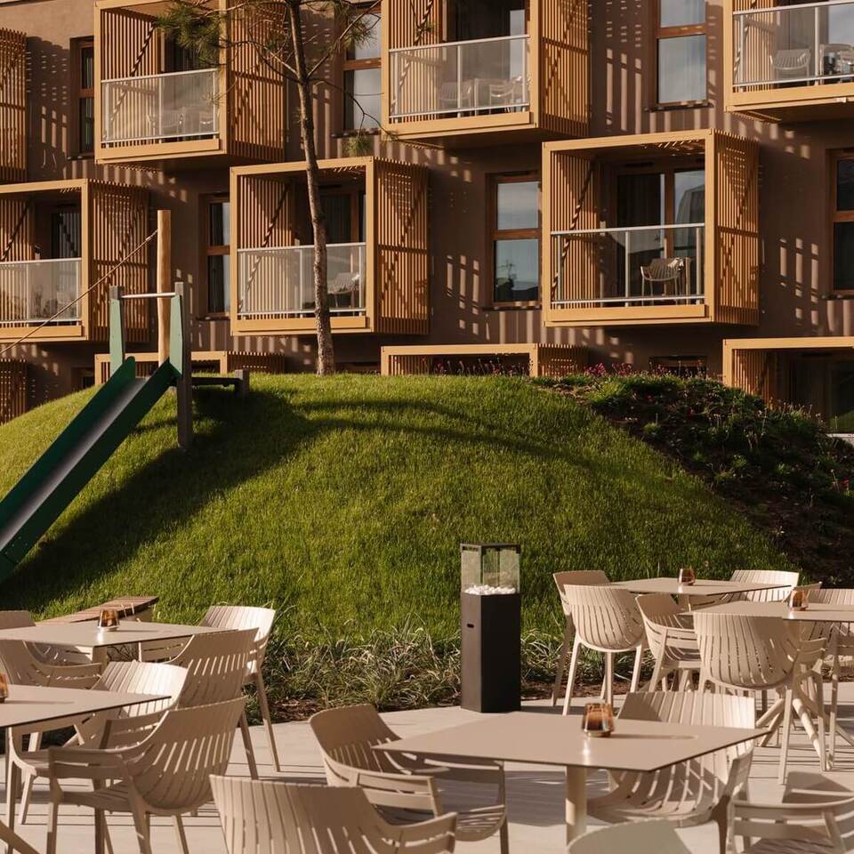 Taras hotelowy z stolikami na świeżym powietrzu, zielony pagórek ze zjeżdżalnią i nowoczesne drewniane balkony z krzesłami.