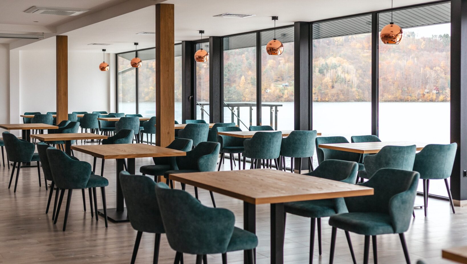 Hotelrestaurant am See mit türkisfarbenen Polsterstühlen, Holztischen, kupfernen Hängelampen und weitem Blick aufs Wasser