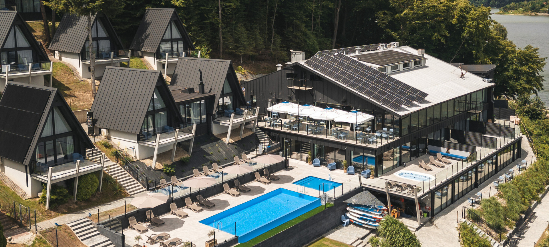 Zakwaterowanie nad jeziorem: domki typu A, budynek z panelami słonecznymi, taras restauracyjny, basen, leżaki i deski SUP.