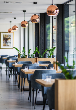 Restauracja hotelowa z drewnianymi stolikami, szarymi krzesłami, miedzianymi lampami i dużymi oknami z widokiem na wodę.