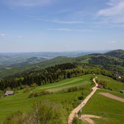 Panoramic mountain vista with rolling green hills, forested ridges, a winding dirt road and scattered cottages under clear blue sky
