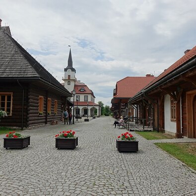 Cobblestone courtyard with wooden lodges, red-tiled roofs, clock tower, benches, flower planters and guests.