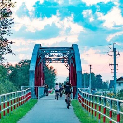 Cyclists riding across a red metal truss bridge at sunset, tree-lined path and nearby houses