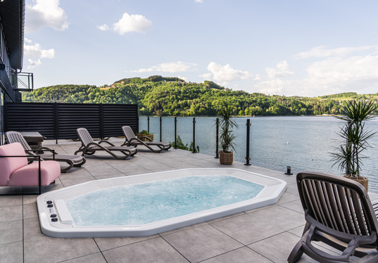 Taras z jacuzzi, leżakami, różowymi fotelami i szklaną balustradą z widokiem na jezioro i zielone wzgórza.