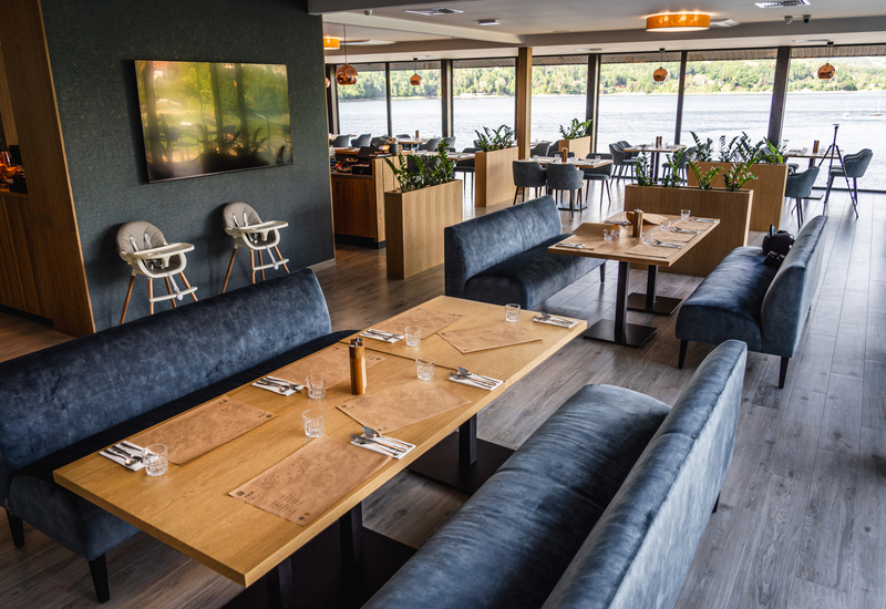 Hotelrestaurant am Wasser mit Seeblick, Holztischen, samtblauen Sitzbänken, Pflanzen und zwei Hochstühlen für Kinder.