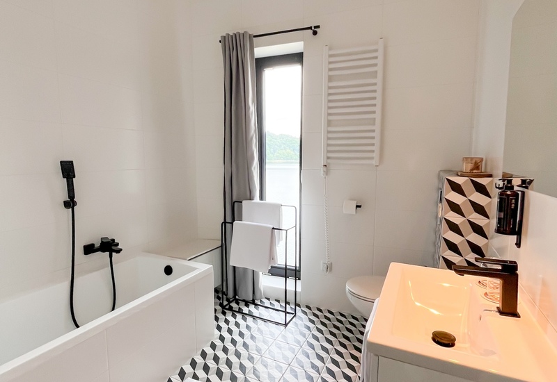 Helles, modernes Hotelbadezimmer mit Badewanne, schwarzer Duscharmatur, geometrischen Bodenfliesen, Handtuchheizkörper, Waschbecken und Fenster